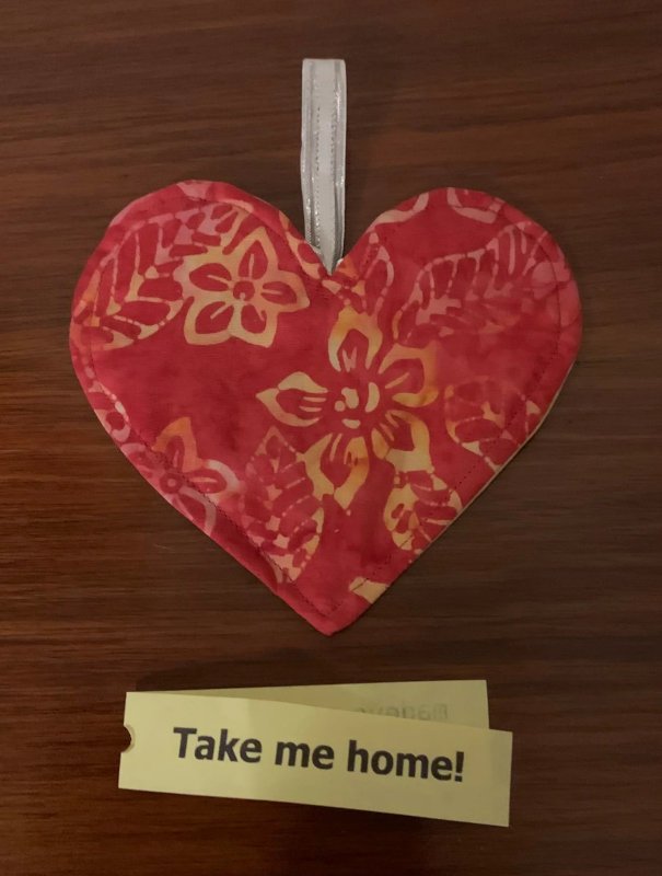 A flat fabric heart on a wooden surface. The fabric is dark pink, with tropical flowers in a yellow and pink batik design. There is a tag under the heart that reads Take Me Home!