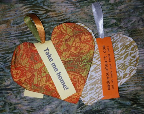 Two flat fabric hearts, overlapping. The one on the right is orange, and has a paper tag attached to it that reads Take Me Home!
The one on the right is mottled gold and white and has a tag that reads Made You A Heart dot com let us know you found one. 
