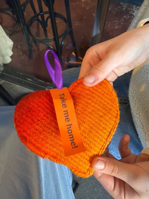 A flat fabric heart being held in a child's hands. The heart is shades of orange in a thick woven fabric and has a purple ribbon at the top for hanging. An orange tag hanging off the ribbon reads Take Me Home.