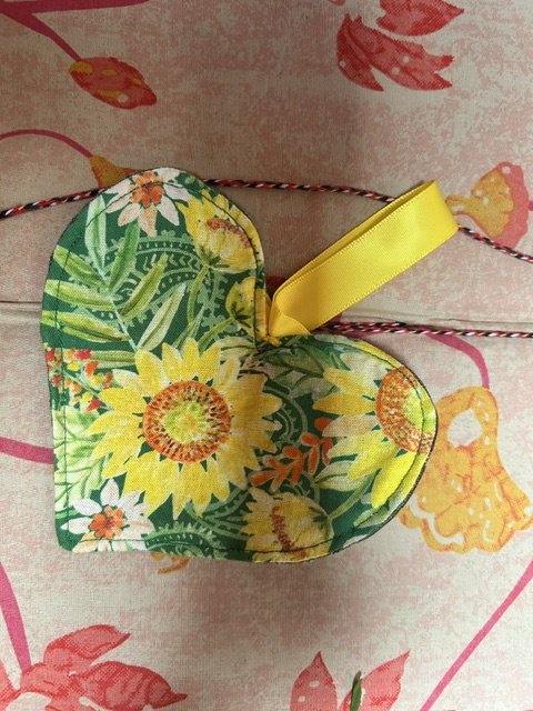 A flat fabric heart resting on a pinkish background. The heart has a yellow ribbon loop to hang it with, and the fabric shows bright yellow sunflowers on a green leafy background.